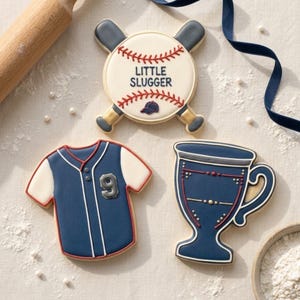 May include: Three decorated sugar cookies shaped like baseball items. One cookie is a baseball with two crossed bats and the text LITTLE SLUGGER. Another is a blue and white baseball jersey with the number 9. The third is a blue trophy cup.
