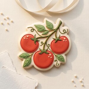 May include: A decorated sugar cookie shaped like a cluster of tomatoes on a vine. The tomatoes are red with white accents, and the leaves are green. The cookie is on a white surface with a ribbon.