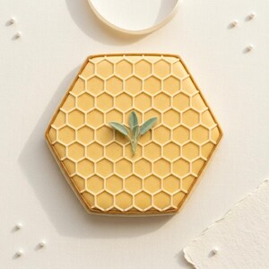 May include: A hexagonal biscuit decorated with a honeycomb pattern and a sprig of green leaves. The biscuit has a yellow base with a lighter yellow honeycomb design and a brown border. A sweet treat.