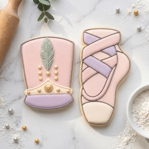 May include: Two decorated sugar cookies shaped like a marching band hat and a ballet slipper. The hat is pink with a light green feather, gold accents, and a purple brim. The ballet slipper is pink and cream with crisscross purple ribbons.