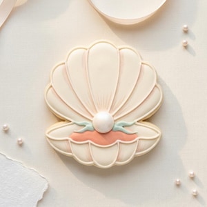 May include: A seashell-shaped cookie with a pearl. The cookie is decorated with white, pink, and light green icing. The pearl is a large, round, white candy. Several small pearl candies are scattered around the cookie on a white surface.