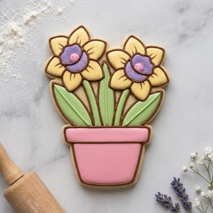 May include: A decorated sugar cookie in the shape of two daffodils in a pink flower pot. The flowers have yellow petals, purple centers, and green leaves. The cookie is outlined in brown.