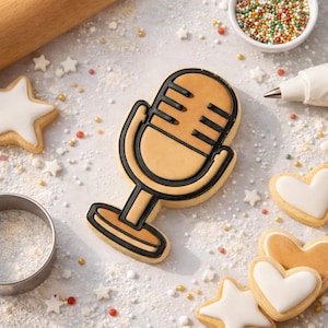 May include: A microphone-shaped cookie with black outline and tan filling, surrounded by star and heart-shaped cookies, sprinkles, and baking tools. The cookies are decorated with white icing. The scene suggests a baking theme.