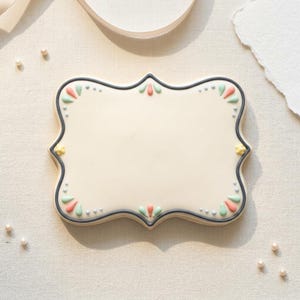May include: A rectangular cookie with a scalloped edge, decorated with pastel-colored floral accents. The cookie has a cream-colored base, a dark gray border, and small pearl-like sprinkles. The design includes yellow, pink, and green details.
