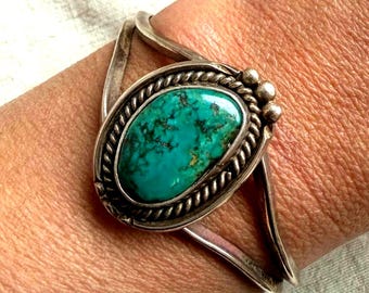 Pode incluir: Pulseira de punho de prata com uma pedra turquesa oval. A turquesa é de uma cor verde-azulado vibrante com padrões de matriz escuros. A pedra é engastada em uma moldura de prata com um design de corda torcida e pequenas contas de prata. A pulseira tem um design simples e aberto.