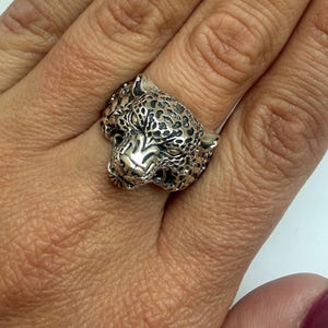 May include: A silver-toned ring featuring a detailed, three-dimensional leopard head design. The metal surface has an oxidized finish to highlight the feline facial features, including the ears, eyes, nose, and the spotted pattern on the head.