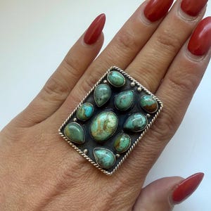 May include: A large, rectangular silver ring featuring multiple turquoise stones in various shapes. The ring has a detailed, rope-like border and a dark background that highlights the vibrant blue-green hues of the stones. The ring is on a hand with red nail polish.