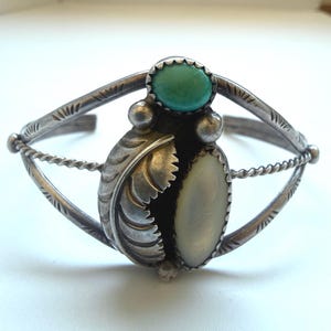 May include: Silver cuff bracelet featuring a central design with a turquoise stone and a mother-of-pearl oval. The bracelet has a detailed feather design and twisted rope accents, showcasing a vintage aesthetic.