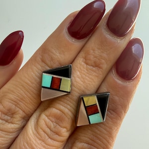 May include: A pair of geometric stud earrings featuring a rhombus shape with inlaid segments of black, turquoise, red, yellow, and iridescent shell. The earrings are framed in a dark metal setting, showcasing a multi-colored mosaic pattern.