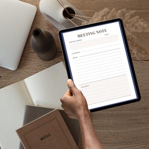 May include: A tablet displaying a "MEETING NOTE" template is held by a person. The tablet is surrounded by notebooks, a laptop, and decorative vases on a wooden surface. The notebook in the foreground has the word "NOTES" on the cover.