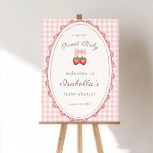 Può includere: Un cartello di benvenuto per un baby shower su un cavalletto di legno. Il cartello presenta uno sfondo a quadretti rosa e una cornice ovale con il testo "A Berry Sweet Baby" e "Benvenuti al baby shower di Isabella". La data è il 28 marzo 2026.