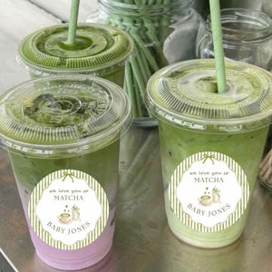 May include: Two clear plastic cups filled with a layered matcha drink, featuring a green top layer and a pinkish-white bottom layer. Each cup has a clear lid and a green paper straw. The cups have a white sticker that says "we love you so MATCHA" and "BABY JONES".