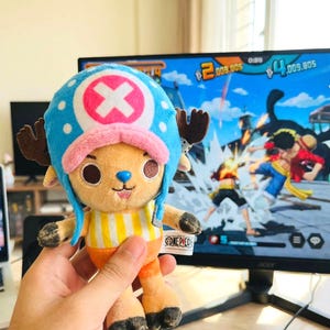 May include: A plush toy of the character Chopper from the anime series One Piece. The toy has a blue hat with white polka dots and a pink cross, brown antlers, and a yellow and orange outfit. The One Piece logo is visible.