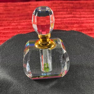 May include: A clear crystal perfume bottle with a faceted, oval-shaped stopper. The bottle has a gold-colored base and a clear, rectangular body. The bottle is sitting on a black surface, with a red textured background.
