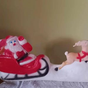 May include: A vintage Christmas decoration showing Santa Claus in a red sleigh pulled by two reindeer. The sleigh and reindeer are white, with red accents on Santa's suit and the reindeer's harnesses. A classic festive scene.