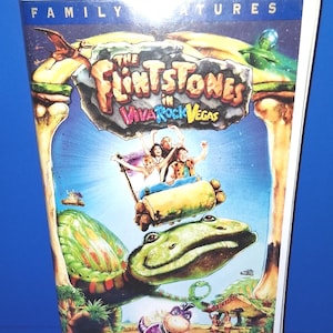 Può includere: Una videocassetta VHS del film "The Flintstones in Viva Rock Vegas". La copertina presenta una scena a cartoni animati colorata con il titolo del film, personaggi sulle montagne russe e un paesaggio a tema dinosauri. Le parole "Family Features" sono in alto.