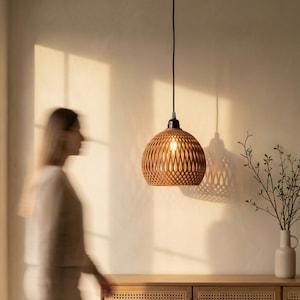 May include: A woven, brown pendant light hangs above a wooden cabinet with woven door panels. The light casts a patterned shadow on the wall. A vase with greenery sits on the cabinet. A person walks past the cabinet.