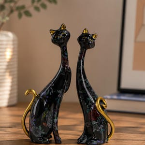 Könnte beinhalten: Zwei schwarze Katzenfiguren mit goldenen Akzenten. Die Katzen haben ein Blumenmuster und goldfarbene Ohren und Schwänze. Die Figuren sind in sitzender Position mit eingerollten Schwänzen dargestellt. Der Hintergrund ist unscharf, was auf eine Wohnkultur hindeutet.