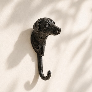 Könnte beinhalten: Ein schwarzer Wandhaken in Form eines Hundekopfes mit Hals. Der Haken erstreckt sich vom unteren Teil des Halses nach unten. Das Objekt hat eine texturierte, dunkle Oberfläche und ein sichtbares Schraubloch zur Befestigung an einer Wand.