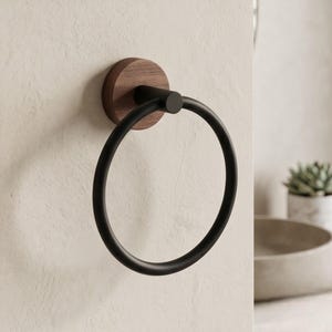May include: A modern towel ring with a matte black finish. The circular ring is mounted on a round, dark wood base. The fixture is attached to a light-colored wall, showcasing a minimalist design.