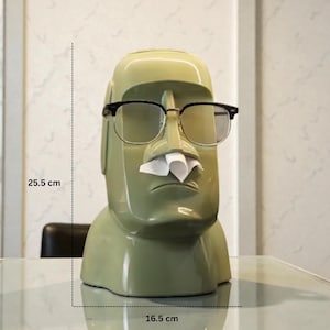 Op de afbeelding: Een groene Moai-vormige tissueboxhouder met een zwart montuur bril en een tissue die uit de neus steekt. De houder is 25,5 cm hoog en 16,5 cm breed. De tissueboxhouder is een uniek en decoratief item.