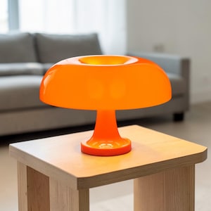 Puede incluir: Una lámpara de mesa naranja con forma de hongo y acabado brillante. La lámpara tiene una pantalla redondeada y una base cilíndrica, colocada sobre una mesita auxiliar de madera clara. Un sofá gris se ve en el fondo.
