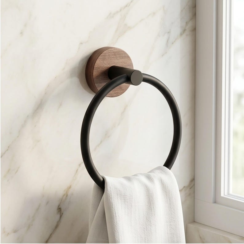May include: A black metal towel ring with a dark wood backing, mounted on a marble-patterned wall. A white towel hangs from the ring. The design is modern and minimalist, suitable for a bathroom.