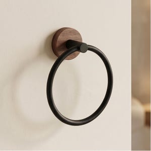 May include: A black metal towel ring mounted on a round, dark wood base. The ring is circular and has a matte finish. The wood base is attached to a white wall, creating a modern bathroom accessory.