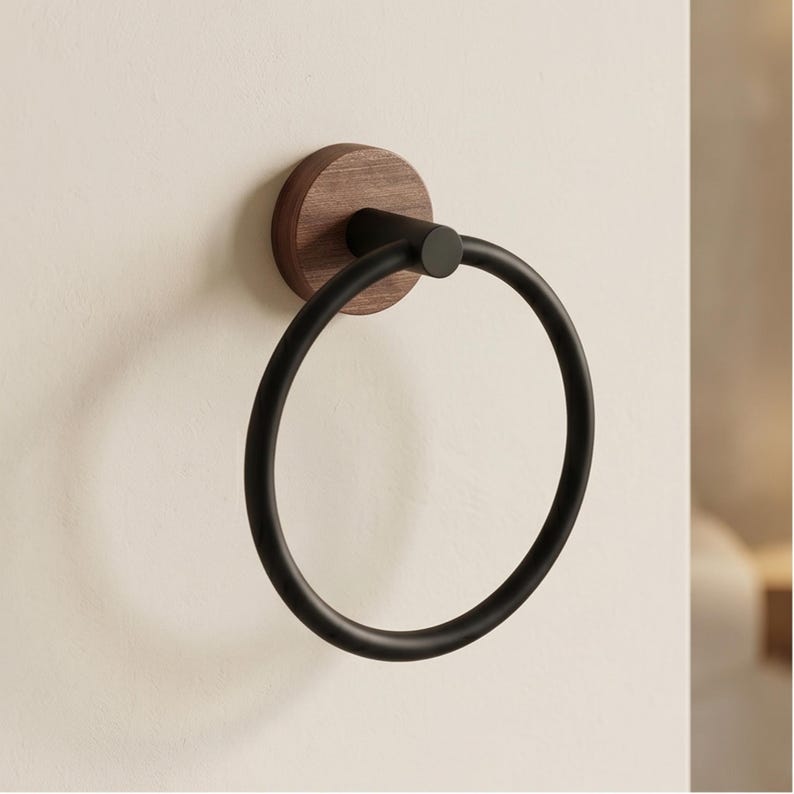 May include: A black metal towel ring mounted on a round, dark wood base. The ring is circular and has a matte finish. The wood base is attached to a white wall, creating a modern bathroom accessory.