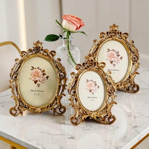 May include: Three ornate, gold-coloured photo frames with oval openings, each displaying a floral print. The frames are 15.2 cm x 20.3 cm and 10.2 cm x 15.2 cm. A vase with a rose is in the background. The frames have decorative scrollwork.