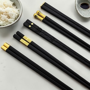 Peut inclure: Des baguettes noires avec des détails dorés sont disposées en diagonale. Les baguettes présentent un design élégant et moderne, avec des détails dorés aux extrémités. Un bol de riz blanc et une petite coupelle avec un liquide foncé sont visibles.