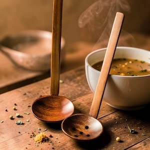 Könnte beinhalten: Zwei hölzerne Suppenkellen mit langen Griffen und runden Schalen, eine mit Abflusslöchern. Eine weiße Schüssel mit dampfender Suppe befindet sich im Hintergrund. Die Kellen und die Schüssel stehen auf einer Holzoberfläche mit Gewürzen.