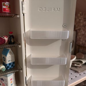 Puede incluir: Un mini-refrigerador blanco roto con la palabra "MAITEC" impresa en la puerta. La puerta tiene tres estantes transparentes. El refrigerador está abierto, revelando estantes con alimentos en miniatura, incluyendo un tarro de mermelada de fresa y un cartón de leche.