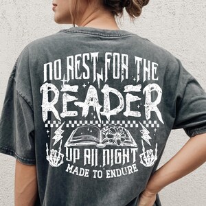 May include: Dark gray t-shirt with white text on the back. The text reads "NO REST FOR THE READER UP ALL NIGHT MADE TO ENDURE" with an open book graphic and lightning bolts. The shirt is worn by a person.