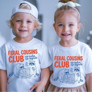May include: A white short-sleeved t-shirt featuring a graphic of two raccoons and the orange and blue text "FERAL COUSINS CLUB no rules no regrets". One raccoon is eating from a trash can while the other holds a snack and a drink.