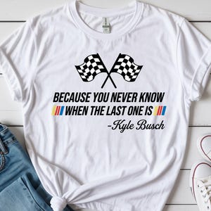 May include: White t-shirt featuring crossed checkered flags and the quote "BECAUSE YOU NEVER KNOW WHEN THE LAST ONE IS" by Kyle Busch. The design includes red, yellow, and blue stripes. The shirt is paired with blue jeans and white sneakers.