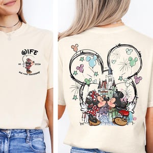 May include: Cream-colored t-shirt with a Mickey and Minnie Mouse design. The front features a small graphic with the word "WIFE" and "Our First Anniversary". The back has a larger design of the couple kissing in front of a castle, inside a Mickey Mouse head outline.