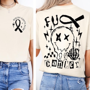 May include: A cream-colored t-shirt featuring a black graphic on the front left chest of a ribbon with the text NOBODY FIGHTS ALONE. The back displays a large black graphic of a melting smiley face, a hand gesture, and the text FUCK CANCER.