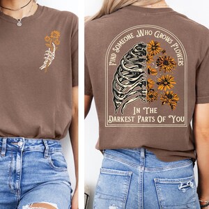 Puede incluir: Una camiseta marr&oacute;n con un peque&ntilde;o gr&aacute;fico en el pecho izquierdo que muestra una mano de esqueleto sosteniendo flores amarillas. La parte posterior muestra un gr&aacute;fico grande de una caja tor&aacute;cica esquel&eacute;tica con flores amarillas y el texto, Find Someone Who Grows Flowers In The Darkest Parts Of You.