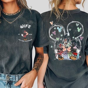 May include: Dark grey t-shirt with a Minnie Mouse design and the text "WIFE" and "OUR FIRST ANNIVERSARY". The back features a Mickey Mouse design with a castle and fireworks. The shirt has a vintage wash finish.