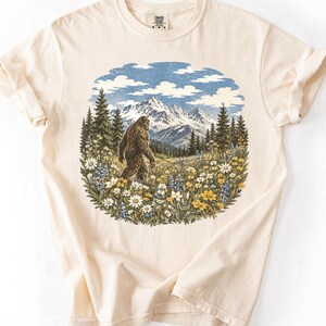 May include: A cream-colored short-sleeve t-shirt featuring a circular graphic print of a brown Bigfoot walking through a field of white and yellow wildflowers, with a backdrop of green pine trees and snow-capped mountains under a blue sky.