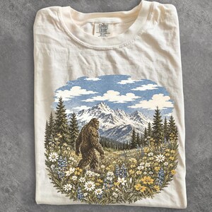 May include: A cream-colored t-shirt featuring a graphic print of a Bigfoot creature walking through a field of wildflowers and pine trees with snow-capped mountains in the background. The inner neck label reads Comfort Colors.