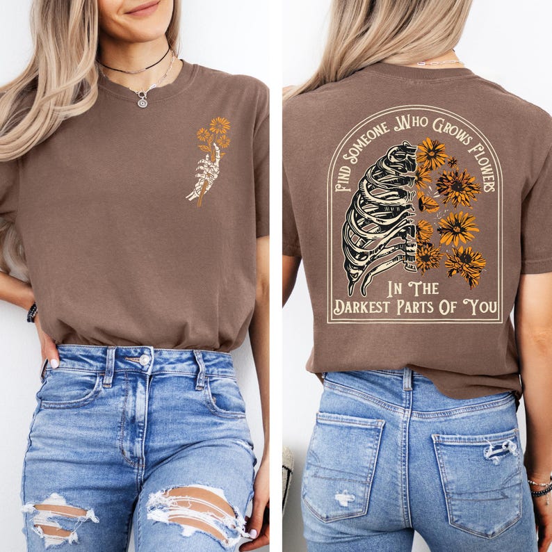 Puede incluir: Una camiseta marr&oacute;n con un peque&ntilde;o gr&aacute;fico en el pecho izquierdo que muestra una mano de esqueleto sosteniendo flores amarillas. La parte posterior muestra un gr&aacute;fico grande de una caja tor&aacute;cica esquel&eacute;tica con flores amarillas y el texto, Find Someone Who Grows Flowers In The Darkest Parts Of You.