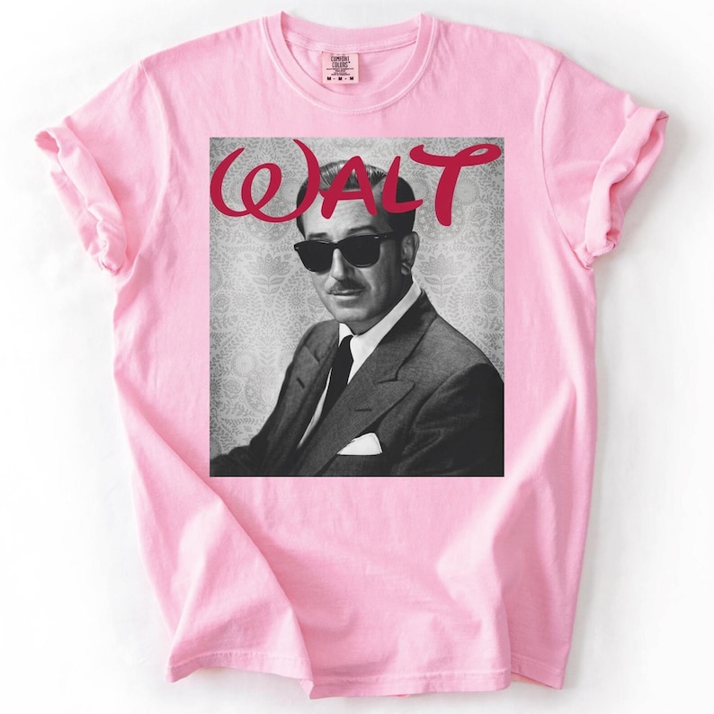 May include: A light pink t-shirt featuring a black and white image of a man wearing sunglasses and a suit. The word "WALT" is written in red cursive above the image. The shirt has short sleeves and a crew neck.