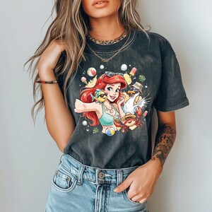 May include: Dark gray t-shirt featuring a colorful graphic of a cartoon character with red hair, a blue top, and sunglasses, taking a selfie with a fish, seagull, and crab. The design includes beach-themed elements like ice cream and a beach ball.