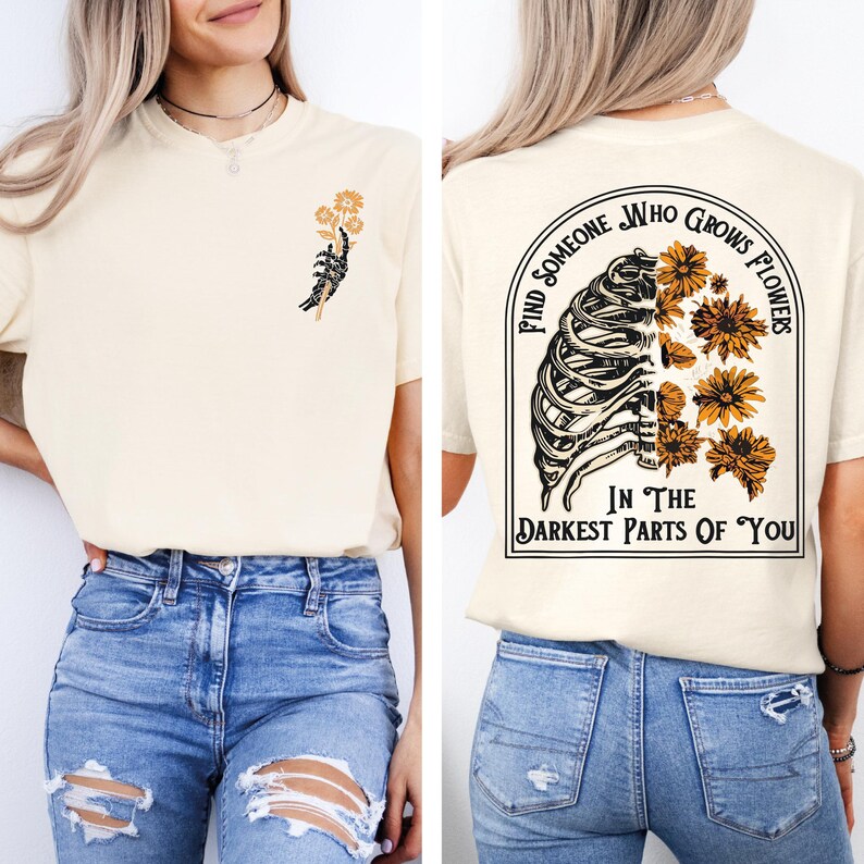 Puede incluir: Una camiseta color crema con un peque&ntilde;o gr&aacute;fico en el pecho izquierdo de una mano de esqueleto sosteniendo una flor amarilla. La parte posterior muestra un gr&aacute;fico grande de una caja tor&aacute;cica fusionada con flores amarillas y el texto Find Someone Who Grows Flowers In The Darkest Parts Of You.