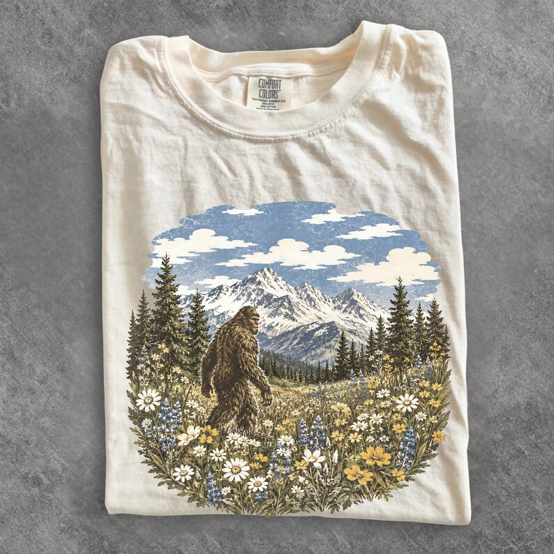 May include: A cream-colored t-shirt featuring a graphic print of a Bigfoot creature walking through a field of wildflowers and pine trees with snow-capped mountains in the background. The inner neck label reads Comfort Colors.