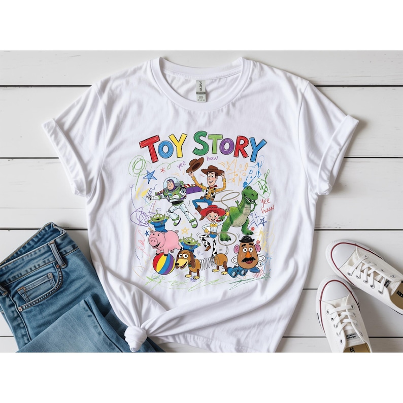 May include: White t-shirt featuring a colorful graphic of Toy Story characters, including Woody, Buzz Lightyear, and Rex. The words "Toy Story" are written in a playful font above the characters. The shirt is paired with blue jeans and white sneakers.