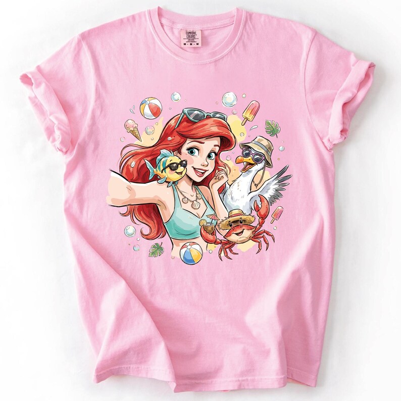 May include: Pink t-shirt featuring a cartoon illustration of a red-haired woman, a yellow fish, a seagull, and a crab, all wearing sunglasses and hats. The woman is taking a selfie, with beach balls, ice cream, and bubbles in the background.