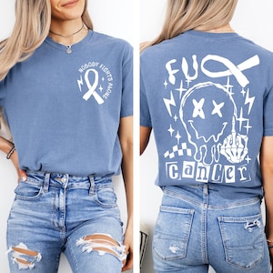 May include: A blue t-shirt featuring a white graphic on the front chest with a ribbon and the text "NOBODY FIGHTS ALONE." The back displays a large white graphic of a melting smiley face, a ribbon, and the text "FUCK CANCER" with a skeleton hand.